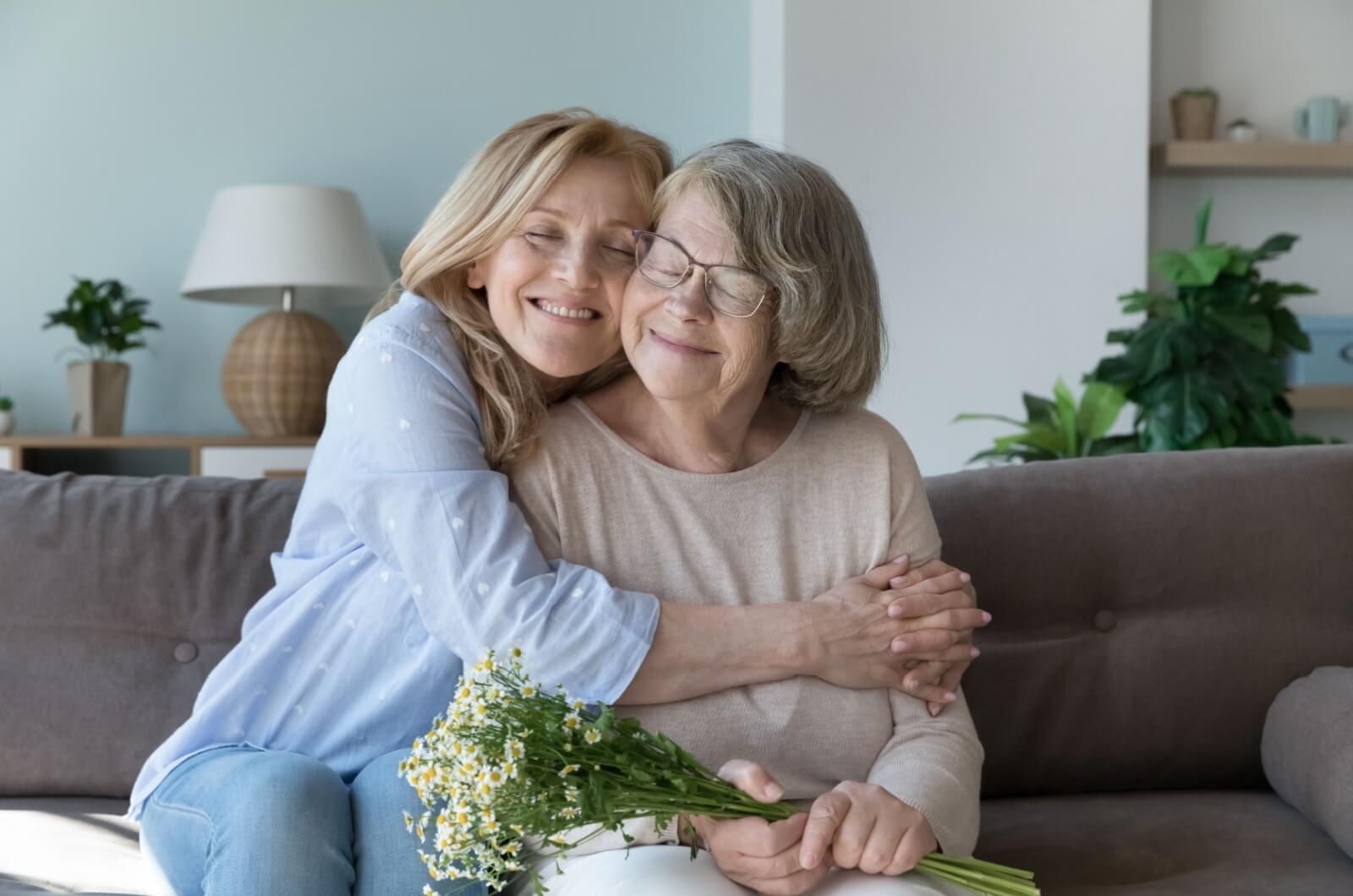 A smiling parent holding flowers while their happy adult child embraces them during a visit in assisted living.